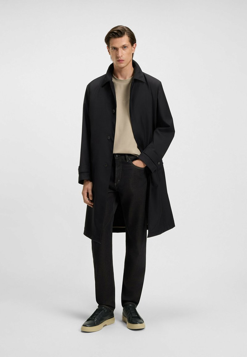 Black trench coat with a smooth finish, button closure, and large collar, worn over a beige shirt and dark jeans with green sneakers.