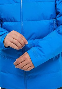 Blaue Puffjacke mit Reißverschluss vorne, verstellbaren Klettverschluss-Manschetten, glatter Textur und gerippten horizontalen Nähten zur Isolierung.