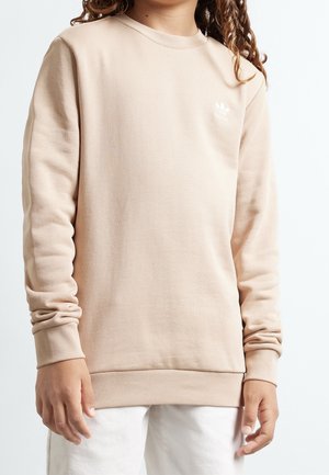 Kind mit langen lockigen Haaren, trägt einen beigen Adidas-Sweatshirt und weiße Hose, steht vor einem schlichten hellen Hintergrund.