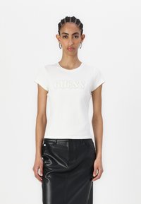 Hvit kortermet t-skjorte med "GUESS" logo i teksturert detaljering. Kombinert med et svart skinnskjørt. Glatt stoff, design med rund hals.