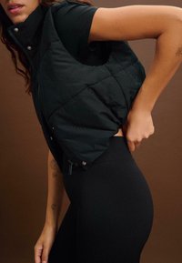 Schwarze Steppweste mit hohem Kragen und Druckknöpfen, getragen über einem schwarzen engen Oberteil und Leggings, die glatte Texturen und ein schlankes Design zur Schau stellt.