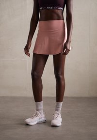 Conjunto deportivo que incluye una camiseta corta oscura y una falda deportiva rosa claro. El calzado consiste en zapatillas blancas y calcetas cortas de color gris claro.