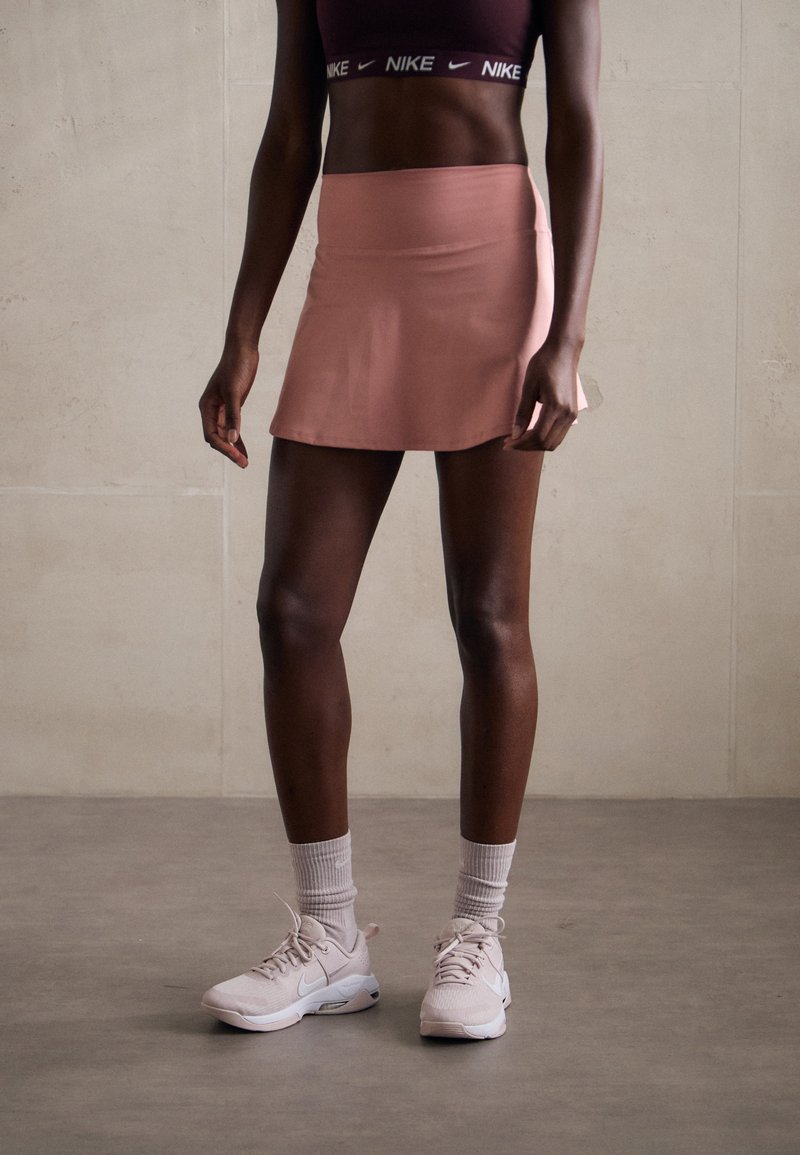 Conjunto deportivo que incluye una camiseta corta oscura y una falda deportiva rosa claro. El calzado consiste en zapatillas blancas y calcetas cortas de color gris claro.