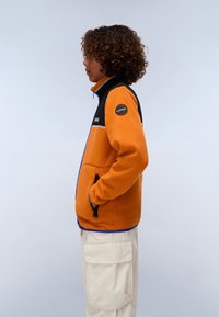 Fleece jakna u narančastoj i crnoj boji, s visokim okovratnikom, džepovima sa zatvaračem i plavim obrubom. Logotip na lijevom ramenu.
