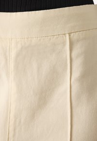 Tessuto beige chiaro con superficie texturizzata. Presenta pieghe cucite e una vita liscia. Si concentra sulla variazione nella texture del tessuto.