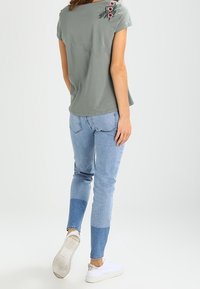 Grå bomulds t-shirt med blomsterbroderi på skulderen, parret med lyseblå denim jeans med farveblokkerede detaljer og hvide sneakers.