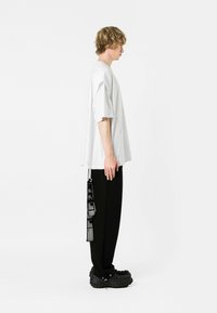 Camiseta blanca de gran tamaño con mangas cortas, combinada con pantalones negros que cuentan con una correa desmontable con texto y una suela texturizada prominente.