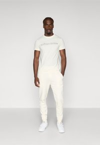 Helles Creme-Farbenes T-Shirt mit kurzen Ärmeln und dem Text "BJÖRN BORG", kombiniert mit passenden cremefarbenen Jogginghosen und weißen Sneakers. Glattes Material, lässige Passform.