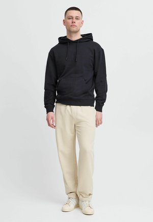 Schwarzer Baumwoll-Hoodie mit Fronttasche, kombiniert mit hellbeigen Jogginghosen. Das Modell trägt weiße Sneakers; lässiges Outfit mit minimalistischem Design.