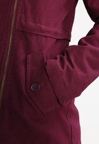 Vinrød jakke med lynlåslukning, sidelomme med knappedetalje og glat tekstureret stof. Fremhævet med et subtilt sømdesign.