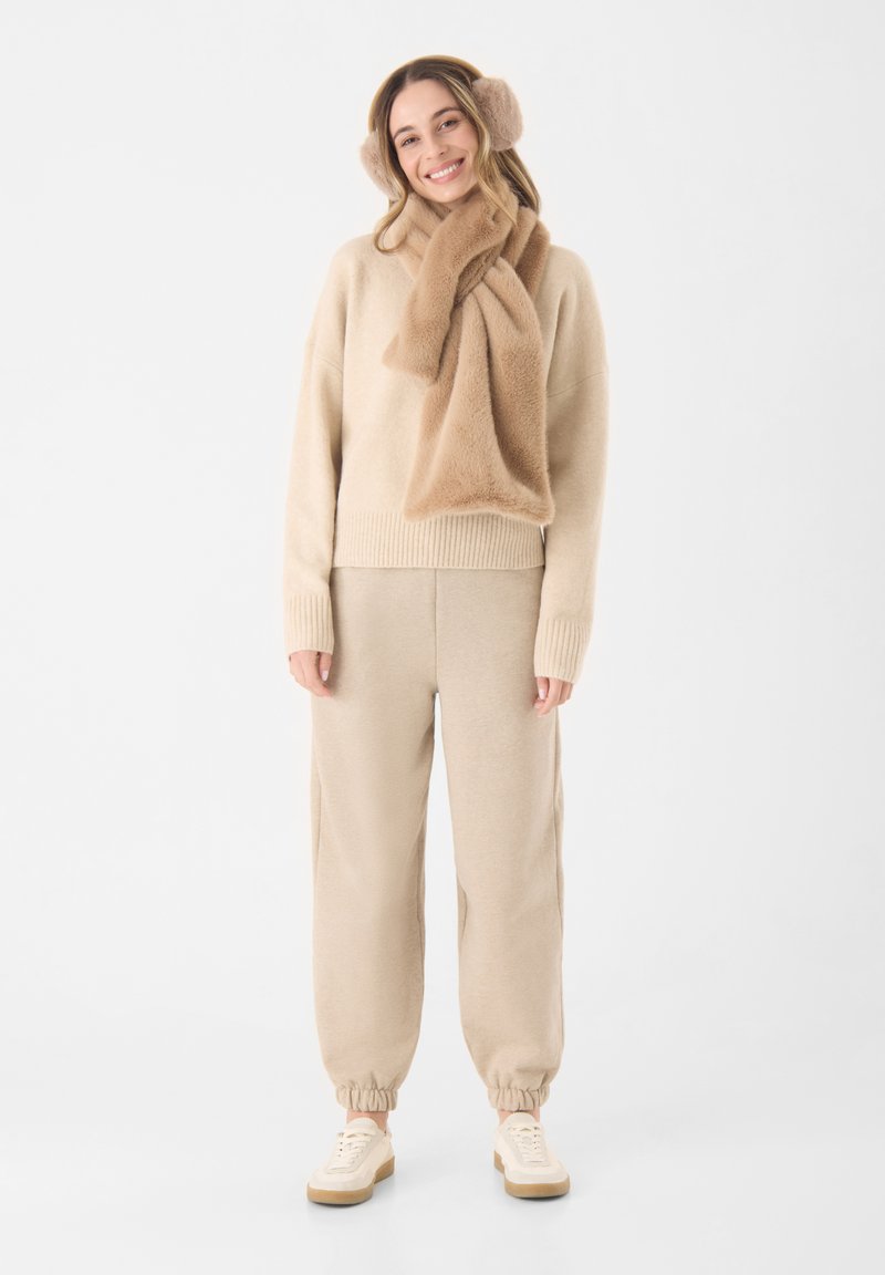 Beige Strickpullover und passende Jogginghose, kombiniert mit einem flauschigen Schal und Ohrenschützern, alles in sanften, neutralen Tönen. Sneakers mit tanfarbenen Akzenten.