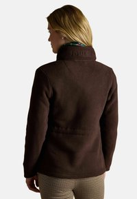 Brązowa polarowa kurtka z wysokim kołnierzem, z wytłoczonym logo "JOULES" z tyłu i gładką teksturą. Noszona z wzorzystymi spodniami.