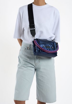 Sac à bandoulière bleu marine en matériau brillant avec des accents tressés violets et roses, équipé d'une sangle noire détachable et d'une fermeture à pression.
