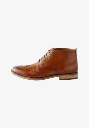 Bottine en cuir marron avec des détails de brogue, design à lacets, bout arrondi et semelle claire contrastante. Fini lisse et texture polie.