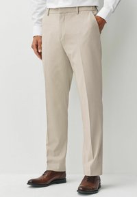 Pantalones de vestir beige con una textura suave, que presentan pasadores para cinturón y un ajuste a medida, combinados con calzado de cuero marrón.