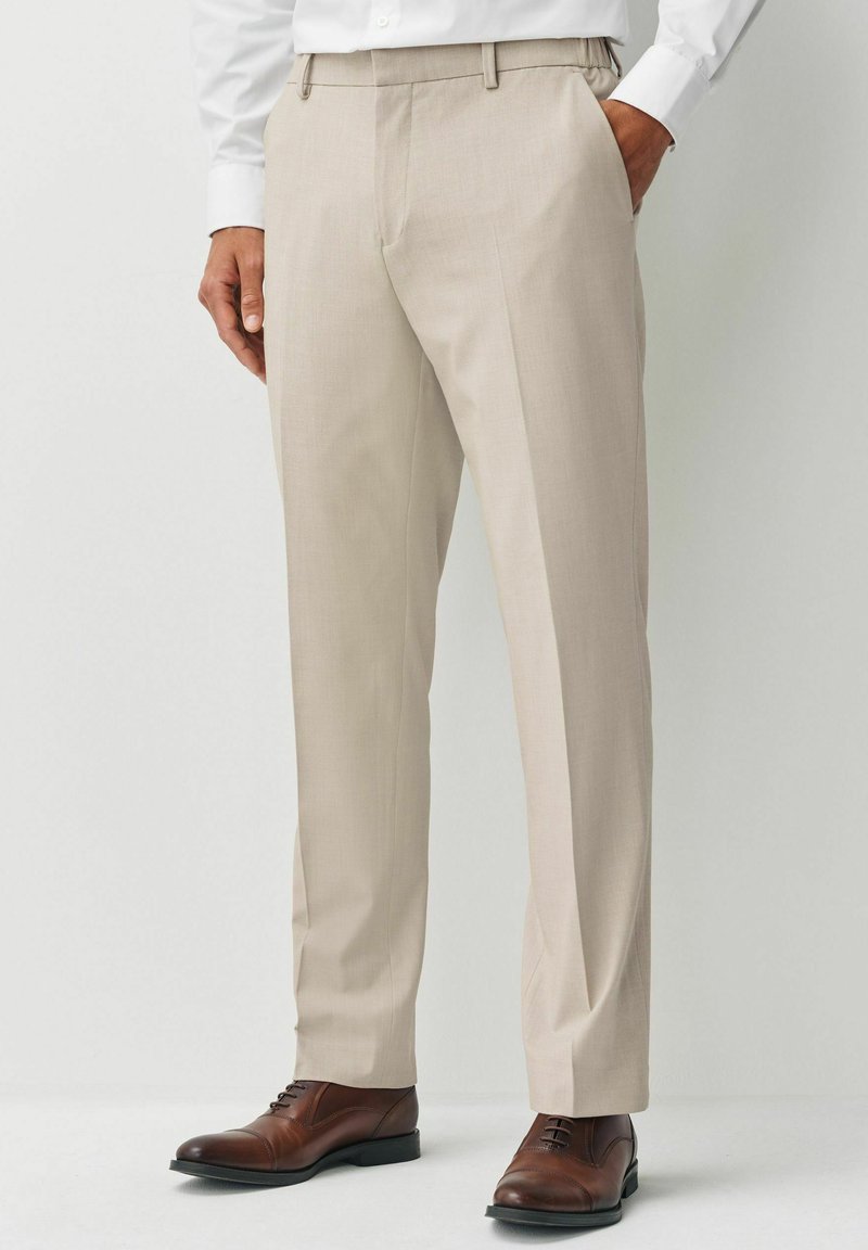 Pantalones de vestir beige con una textura suave, que presentan pasadores para cinturón y un ajuste a medida, combinados con calzado de cuero marrón.