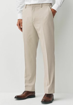 Béžové nohavice s hladkou textúrou, s pútkami na opasok a strihom na mieru, doplnené hnedými koženými topánkami.