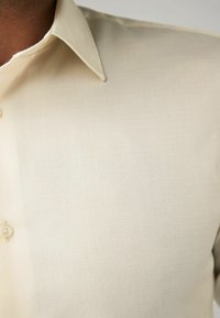 Chemise button-up beige clair, texturée, avec un col distinctif et un motif de losanges subtil, présentant un détail de bouton unique.