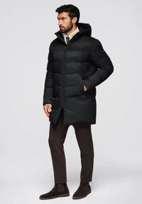 Piumino nero con cappuccio, caratterizzato da una texture trapuntata, zip frontale e tasche laterali. Indossato sopra pantaloni marroni e stivali scamosciati scuri.