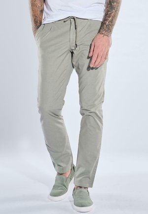 Man wearing light gray drawstring pants, green slip-on shoes, and white T-shirt, standing with hands in pockets against plain background.