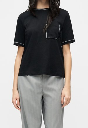Vrouw die een zwart T-shirt met korte mouwen draagt met witte bies en zakdetail, gecombineerd met lichtgrijze broek met fijne strepen.