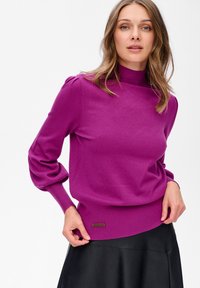 Kobieta w fioletowym swetrze z długimi rękawami i bufiastymi ramionami, w czarnej skórzanej spódnicy, trzymająca krawędź swetra.