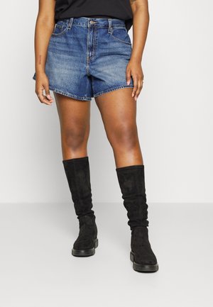 Personne portant un short en denim bleu moyen et des bottes montantes en daim noir, debout devant un fond clair uni.