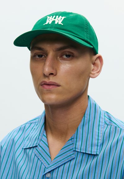 Πράσινο καπέλο με λευκό λογότυπο, κατασκευασμένο από ύφασμα. Το μοντέλο φοράει πουκάμισο με μπλε και πράσινες ριγέ, με μυτερό γιακά και κλείσιμο με κουμπιά.