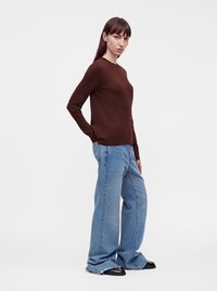 Femme debout, mains dans les poches, portant un haut marron à manches longues, un jean bleu clair ample et des chaussures noires sur un fond blanc uni.