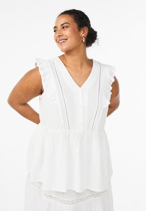 Usmievavá žena v bielej bezrukávovej blúzke s volánmi, zlaté kruhové náušnice, pozerá na stranu s rukami za chrbtom, biele pozadie.