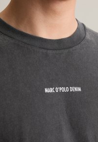 Tmavě šedé tričko s kulatým výstřihem, na němž je bíle vytištěno "MARC O'POLO DENIM". Měkká textura a žebrovaný límec.