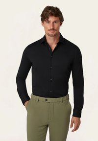 Schwarzes Langarmhemd mit button-down Kragen, kombiniert mit olivgrünen Hosen. Der Stoff wirkt glatt und enganliegend.