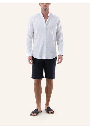 Mann, der ein weißes, langärmeliges Hemd mit Knöpfen, schwarze knielange Shorts und schwarze Sandalen mit offenen Zehen trägt, steht vor einem schlichten Hintergrund.