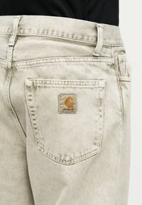 Beżowa tylna kieszeń dżinsów z denimem z łatką z logo Carhartt, noszona z czarnym topem widocznym na wysokości pasa.