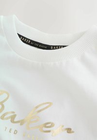Sudadera blanca con cuello acanalado. La parte delantera muestra un logo dorado en cursiva que dice "Baker", con "Ted Baker" en un texto más pequeño debajo.