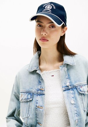 Lyseblå denimjakke med to forlommer, parret med en hvid tekstureret top. En marineblå kasket med hvide accenter og logo fuldender looket.