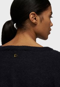 Maglione lavorato a maglia blu navy con scollatura ampia, caratterizzato da un piccolo dettaglio in metallo dorato sulla schiena che mostra il numero "10". Texture morbida visibile.