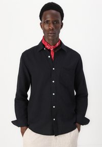 Camicia nera con bottoni, dotata di una singola tasca sul petto, realizzata in tessuto leggero, abbinata a una sciarpa al collo in bandana rossa.