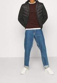 Svart och grå vadderad jacka med huva över en vinröd tröja, kombinerad med ljusblåt löst sittande jeans och vita sneakers.