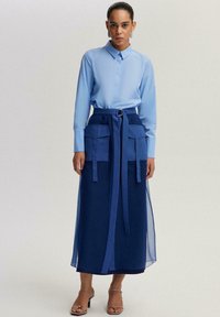 Naine seisab, kandes helesinist nööbitavat särk, pikka tumelillat seelikut suurte taskute ja vööga ning sandaale kontsadel, millel on rihmad.