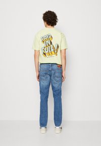 Ljusgul t-shirt med en djärv "BETALD I SIN HELHET!" grafik på baksidan, i kombination med blå denimjeans. Avslappnad passform och mjuk textur.