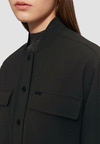 Czarna kurtka bomberka z prążkowanym kołnierzem, zapięciem na guziki i dwiema kieszeniami na piersi. Materiał ma gładką fakturę z lekkim połyskiem.