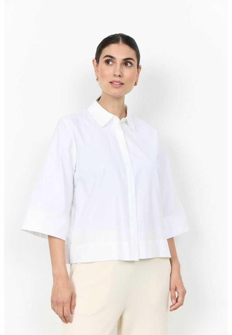 Femme aux cheveux foncés attachés, portant une chemise blanche à manches larges et un pantalon beige clair, regardant légèrement vers le haut et sur le côté.