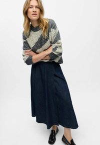 Kvinde med langt blond hår iført en grå og hvid mønstret sweater, mørkeblå midi-nederdel og sorte loafers, poserer med armene krydset.