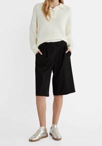 Suéter de punto color crema con cuello, combinado con pantalones cortos negros holgados y zapatillas plateadas metálicas. El conjunto presenta un corte relajado y un diseño casual.