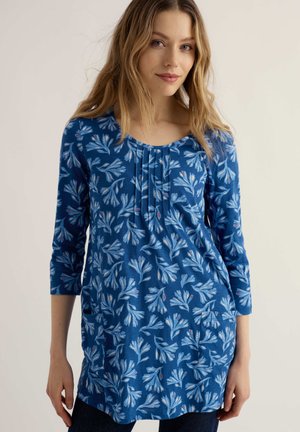 Nő, aki kék, virágmintás, hosszú ujjú tunikát visel elülső zsebekkel, egyszínű, világos háttér előtt.