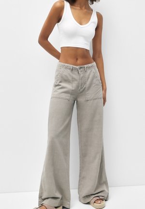 Vrouw die een wit mouwloos crop top draagt, beige wijde broek met hoge taille en open sandalen, staand tegen een effen achtergrond.