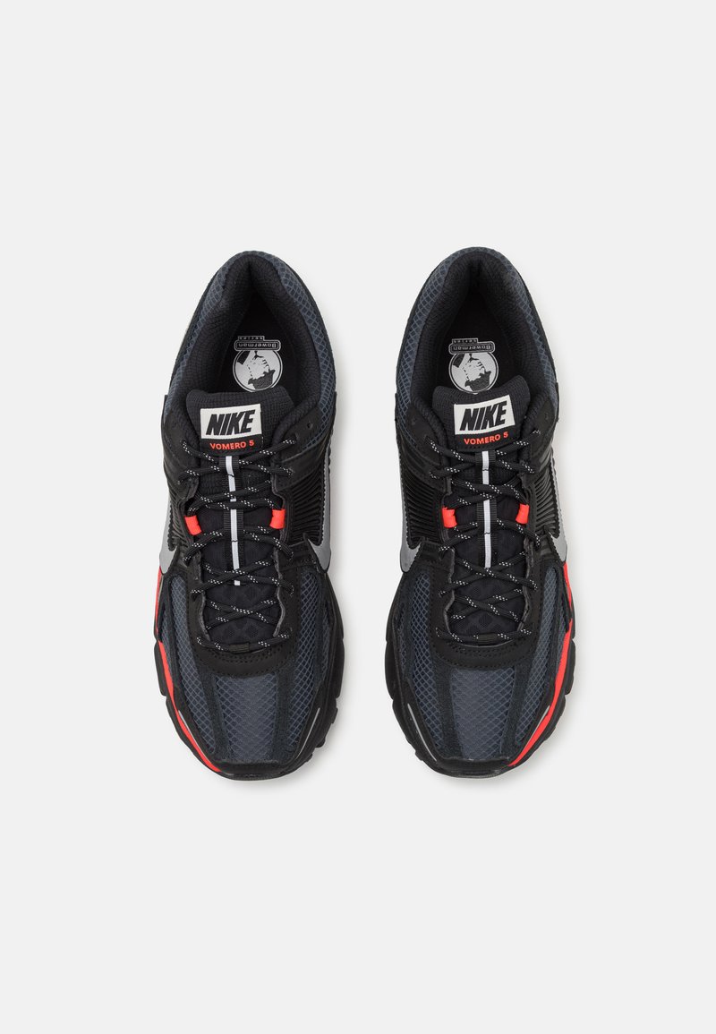 Baskets Nike Vomero 5 avec une tige en mesh noir et gris, des accents réfléchissants, des touches de rouge et une semelle rembourrée pour le soutien.