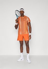 Tenis oblačilo, ki vključuje kratkosleeven svileni majico z oranžnim vzorcem in oranžne kratke hlače, dopolnjeno z belim trakom za glavo in zapestnico.