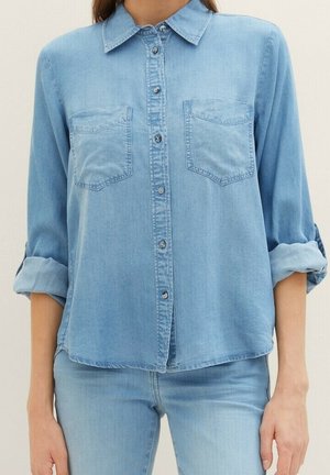 Mujer que lleva una camisa vaquera azul claro con botones, dos bolsillos en el pecho y las mangas remangadas, combinada con jeans azul claro.